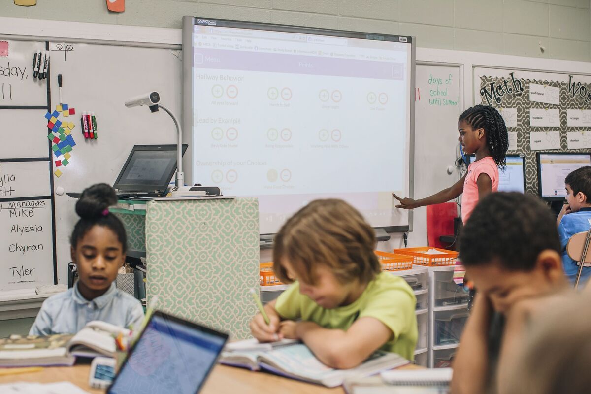 Students work at a table in the foreground, while a student is shown choosing a point from the behavior rubric on a smartboard in the background