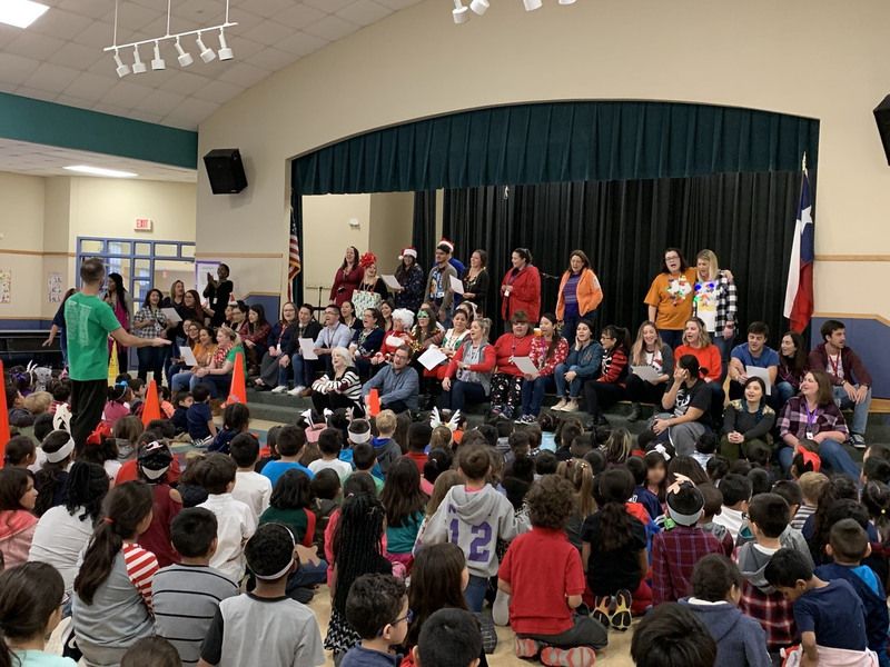 The school gathers at a holiday assembly, showing staff on stage singing and students singing along